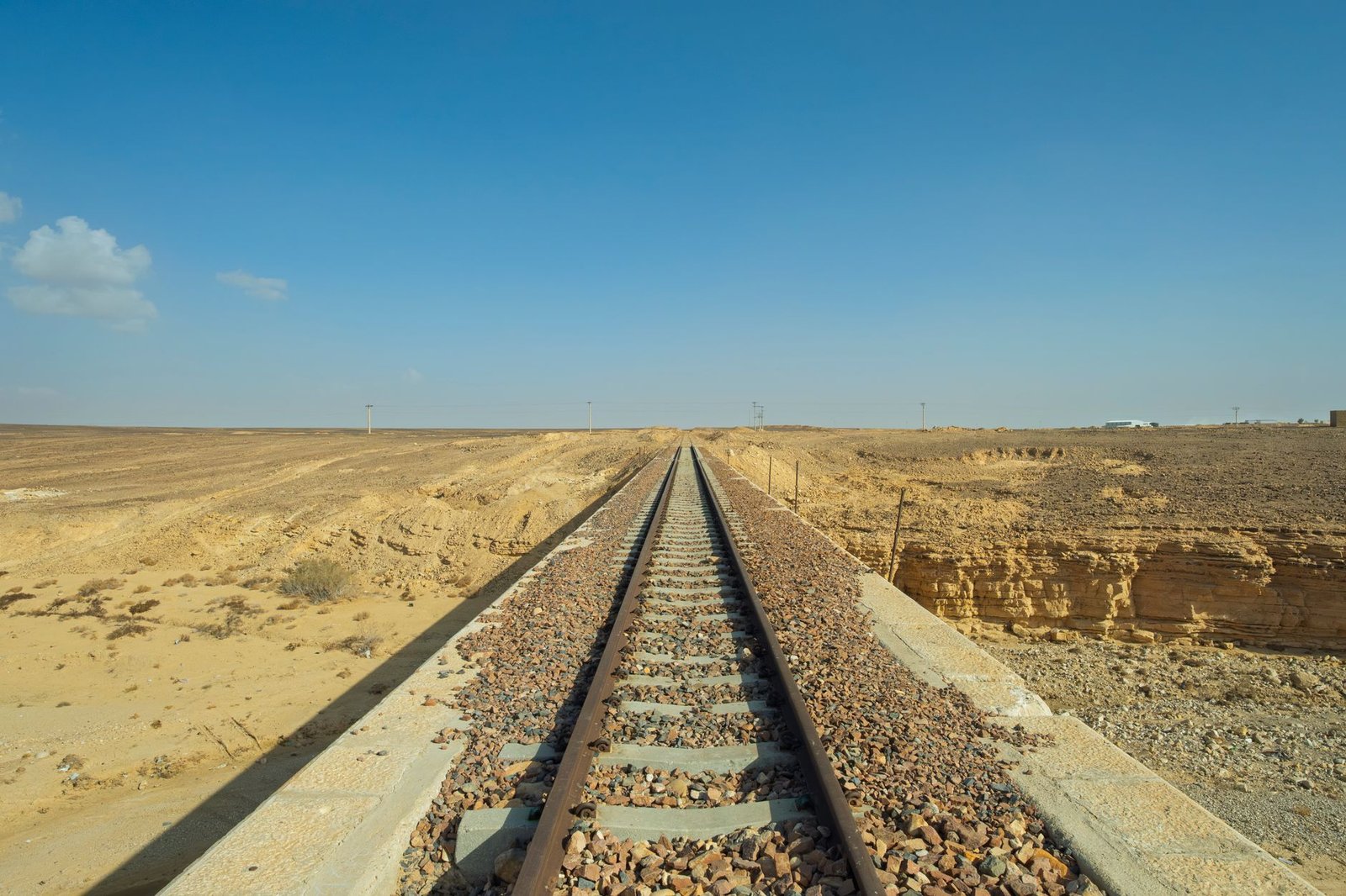 Linha de trem reta sobre brita, atravessando uma planície de deserto seca sob céu azul, com postes baixos ao longo do horizonte.