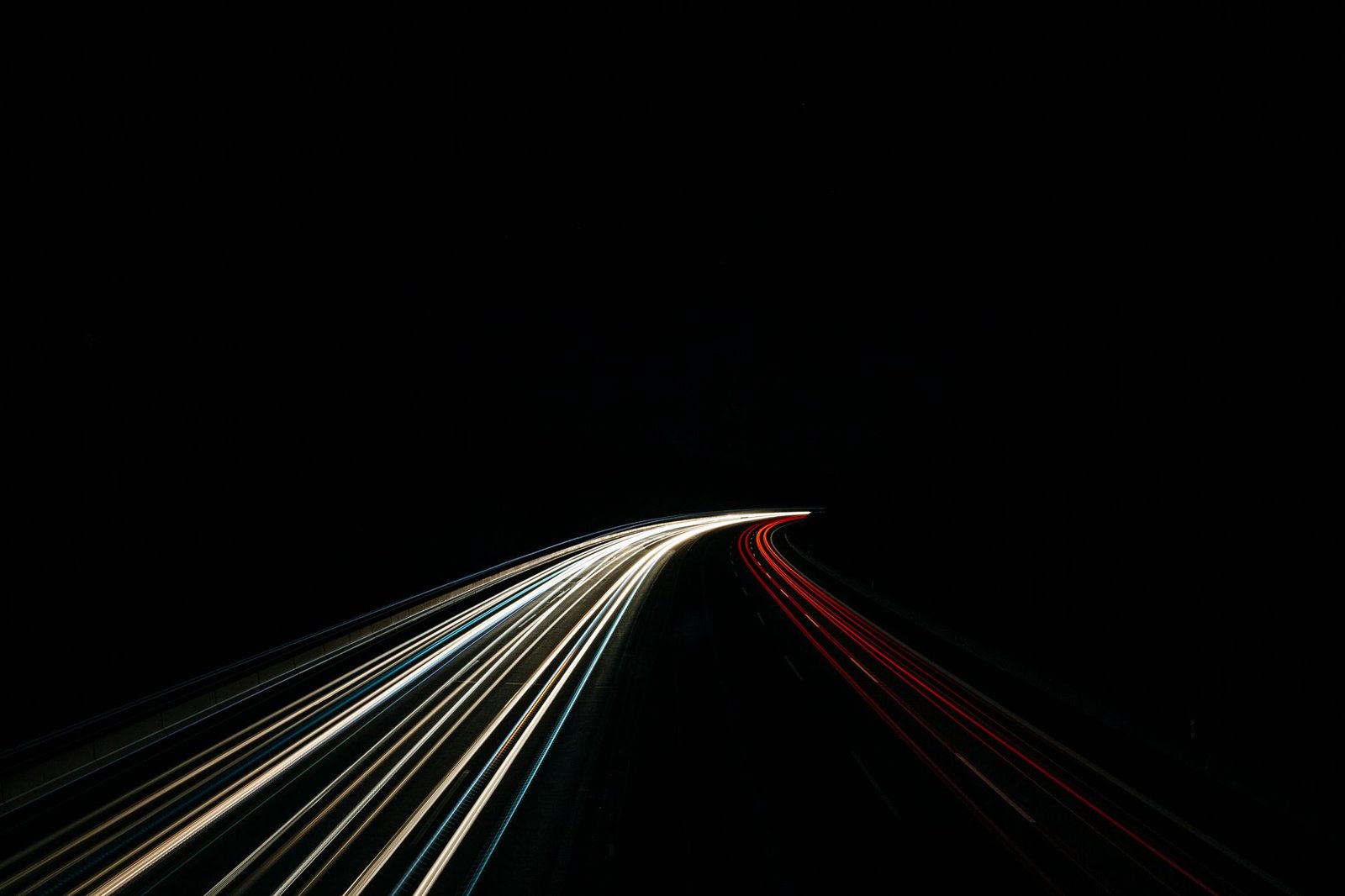 Foto noturna de longa exposição de uma rodovia em curva, com rastros de luz brancos dos faróis de um lado e vermelhos das lanternas do outro