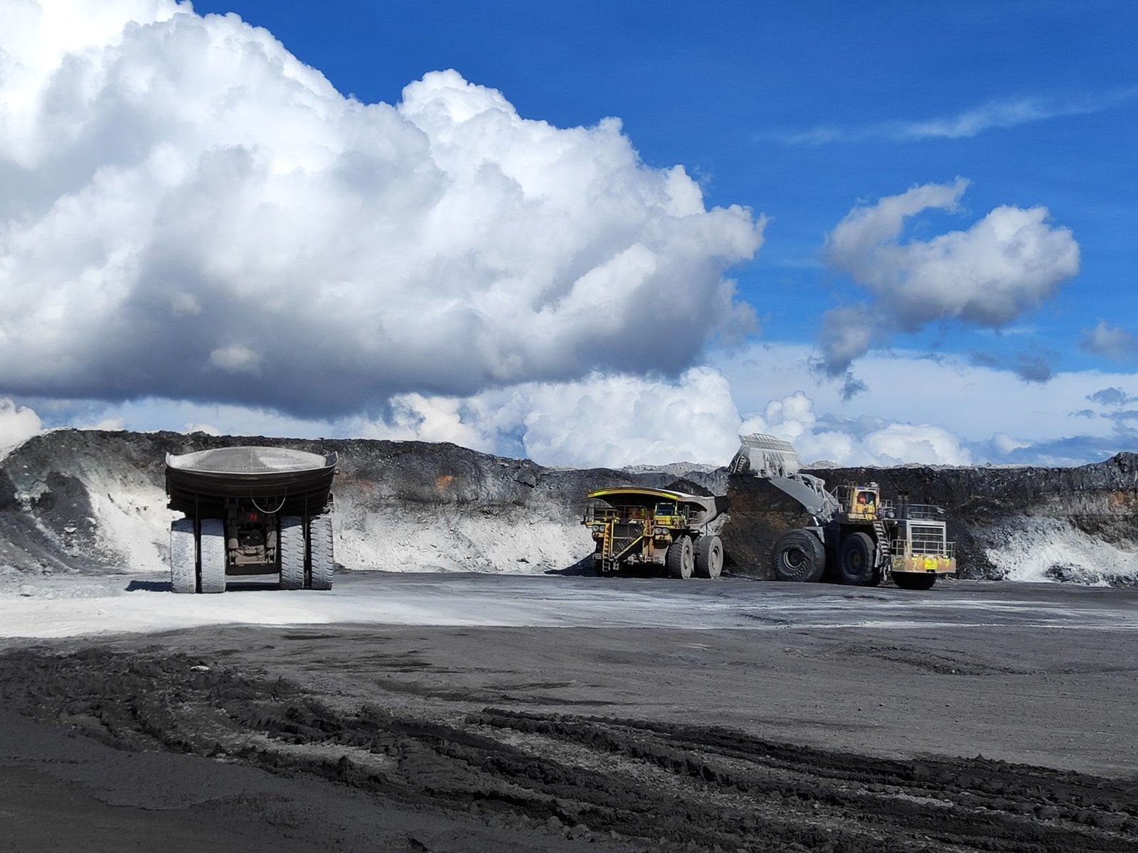 Dois caminhões gigantes de mineração numa mina a céu aberto; uma pá carregadeira enche um deles, sob céu azul com nuvens brancas.