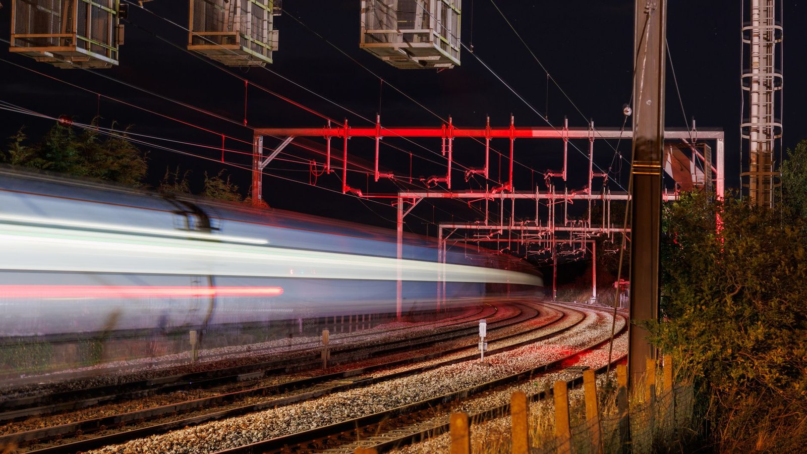 Trem em movimento, borrado pela longa exposição, passa à noite sob fios elétricos e estruturas iluminadas em vermelho, ao lado de trilhos curvos.