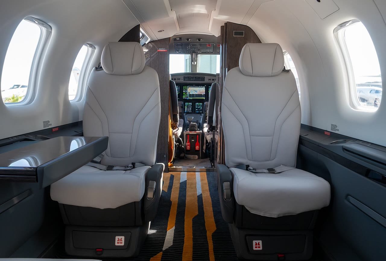 Cabine de passageiros de um Pilatus PC-12 PRO, com duas poltronas de couro claro, uma mesa lateral e, ao fundo, o painel de instrumentos com telas digitais