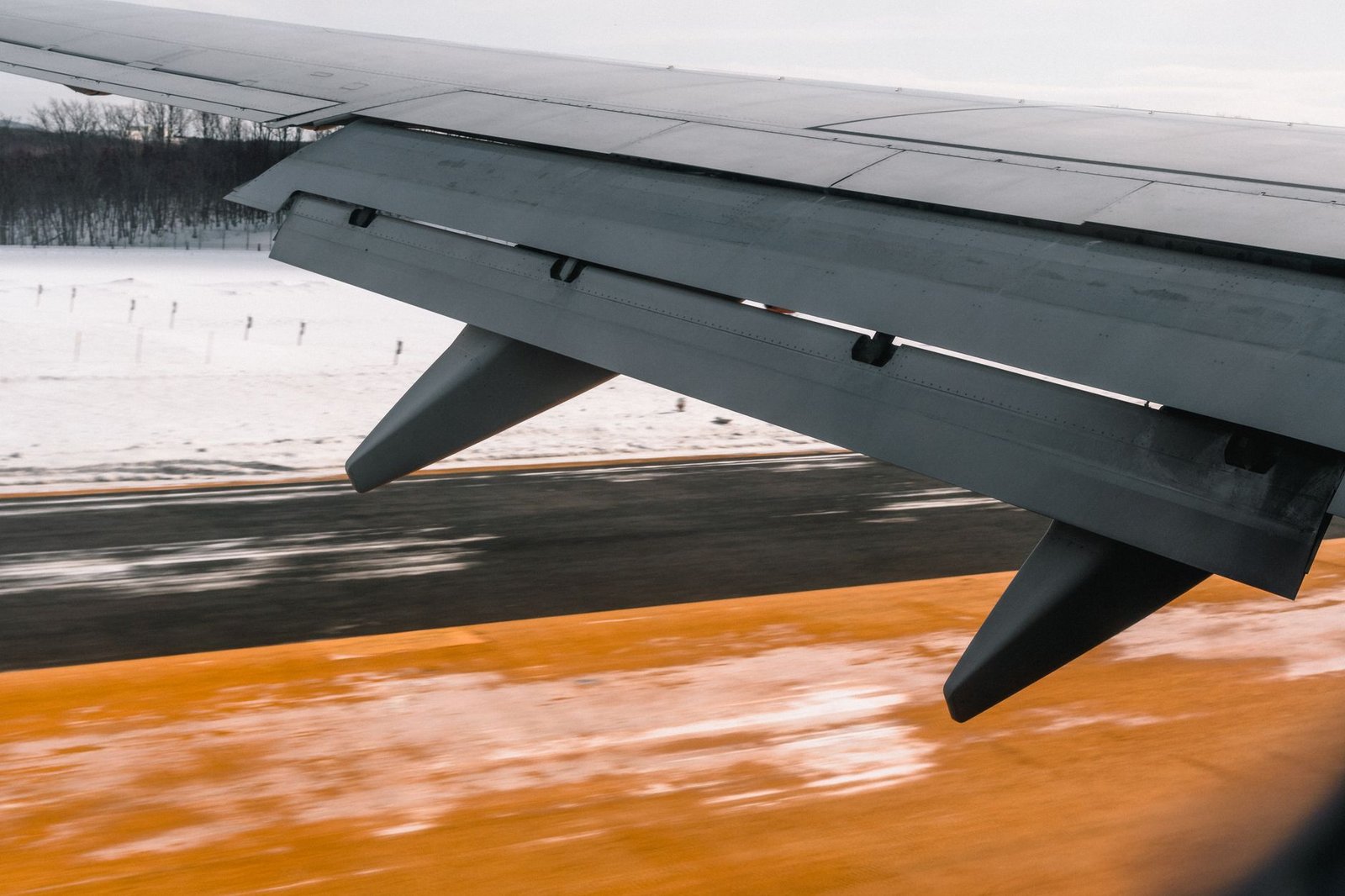 Asa de um avião vista pela janela durante o pouso, com os flapes abaixados sobre a pista e o campo coberto de neve ao lado.