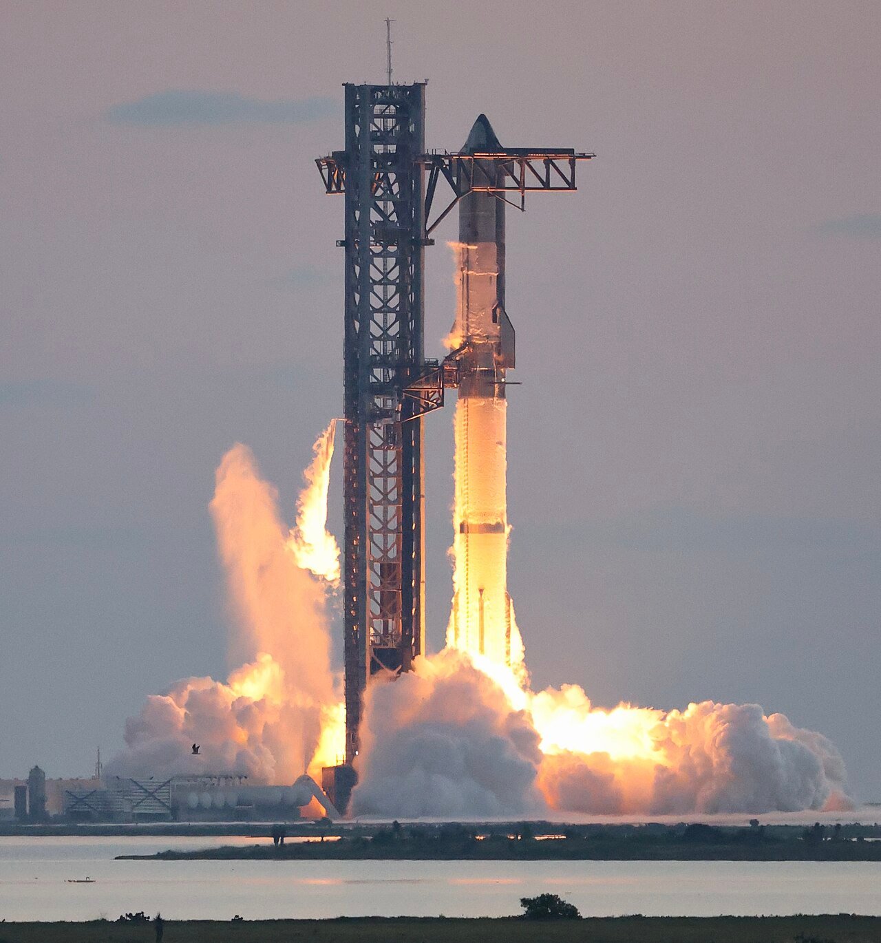 Foguete Starship, da SpaceX, decolando ao lado da torre de lançamento, com uma grande nuvem de fumaça e água em primeiro plano, sob céu nublado, com luz dourada