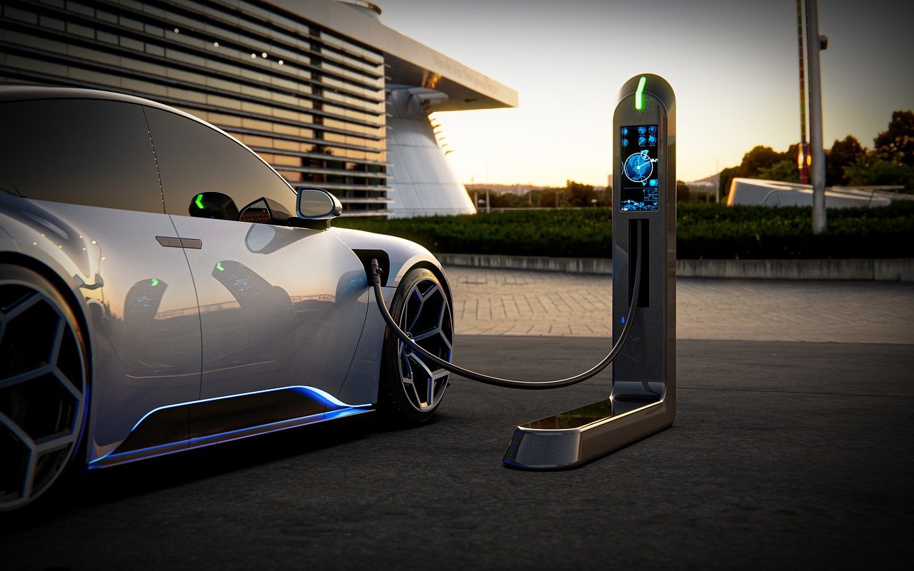 Ilustração em 3D de um carro esportivo prateado ligado por cabo a um carregador moderno com tela iluminada, ao entardecer, diante de um prédio de linhas horizontais.
